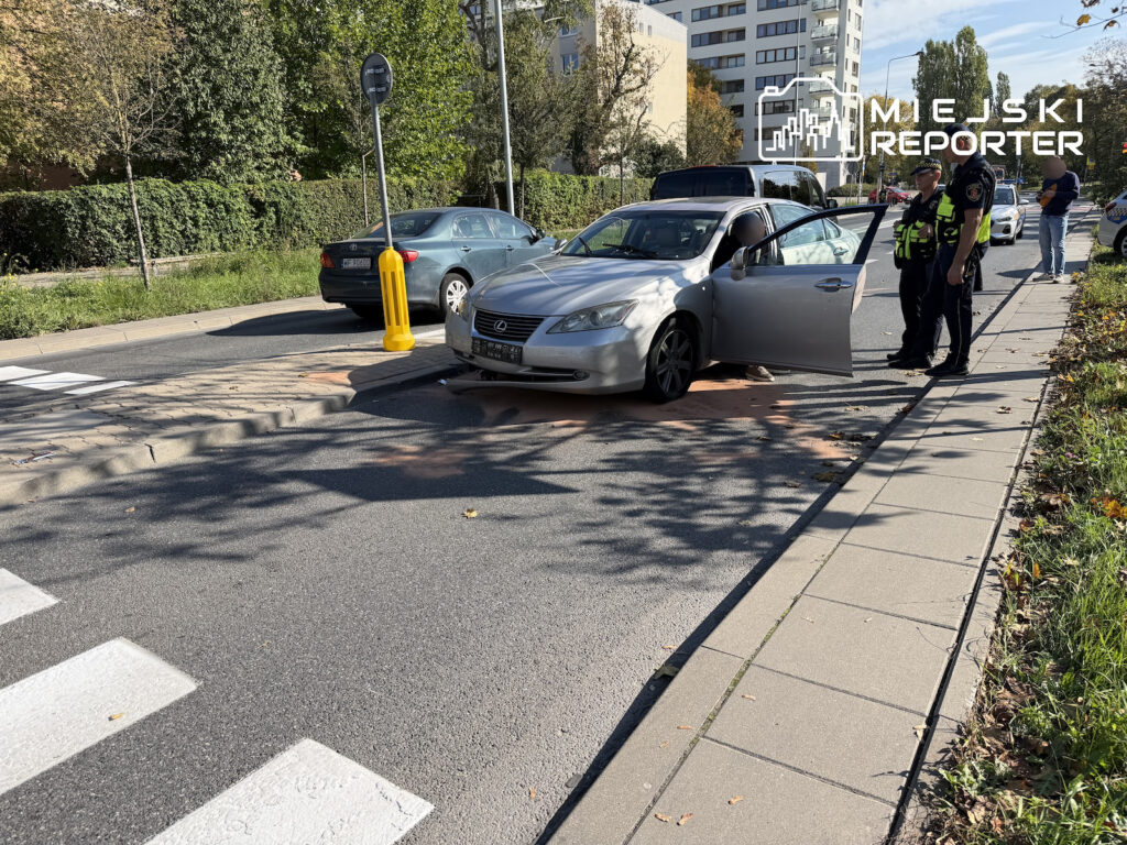 Srebrny Lexus z otwartymi drzwiami stoi na jezdni przy przejściu dla pieszych, obok niego rozmawiają dwaj policjanci.