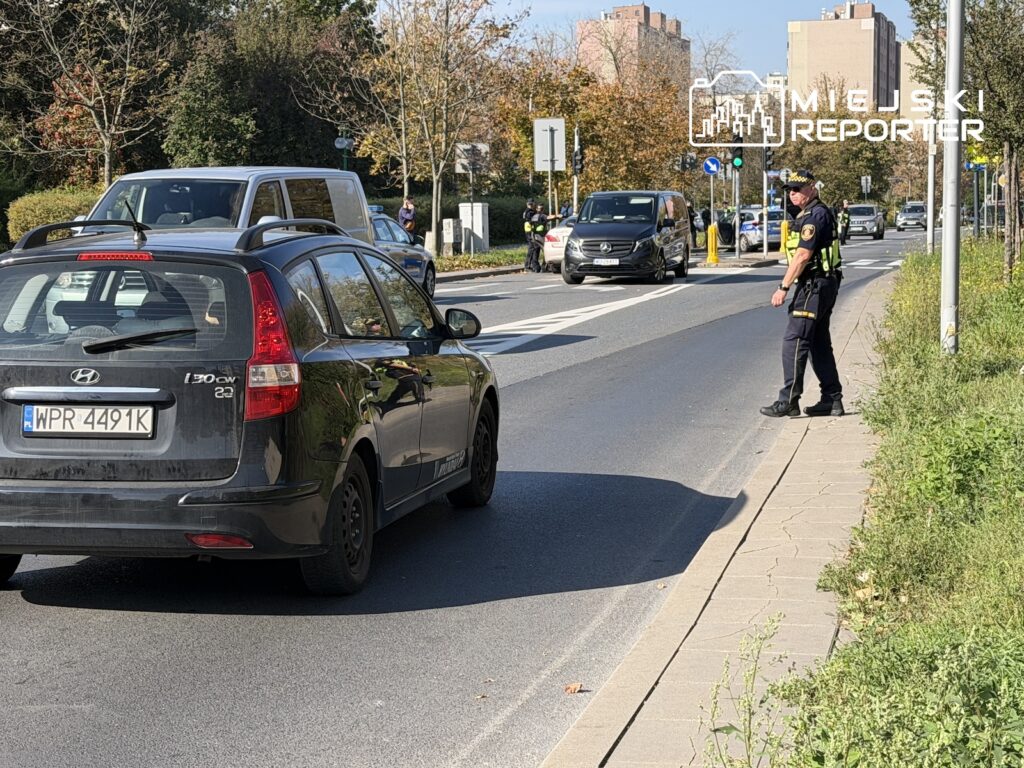 Policjant kieruje ruchem na ulicy, obok stoją samochody i radiowóz.