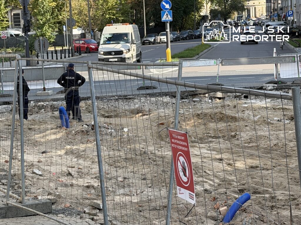 Policjanci stoją przy ogrodzonym wykopie na miejskiej ulicy, obok znaku zakazu wstępu i przejeżdżających samochodów.
