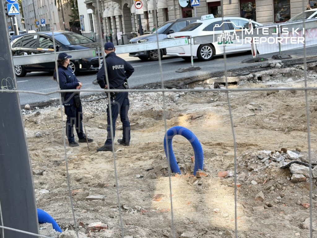 Dwóch policjantów stoi przy ogrodzonym wykopie na rozkopanej ulicy, obok rur i zaparkowanych samochodów.