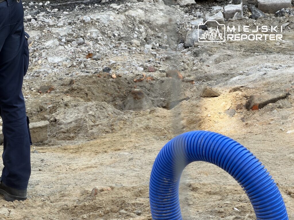 Niebieski karbowany wąż leży wśród ziemi i gruzu na placu budowy, obok stoi osoba w granatowym uniformie.