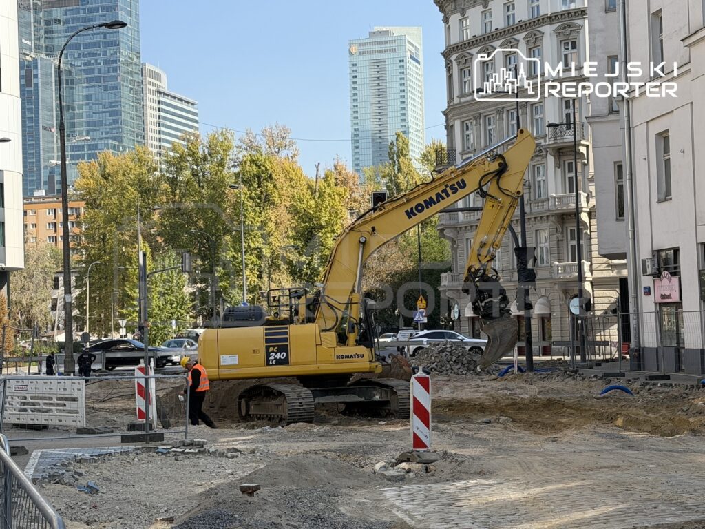 Żółta koparka pracuje na rozkopanej ulicy w centrum miasta, obok niej stoi pracownik w kamizelce odblaskowej.