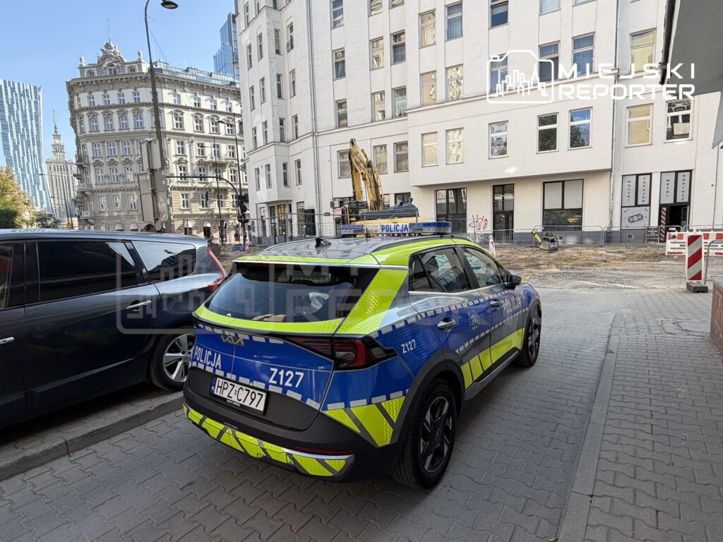 Oznakowany radiowóz policji stoi na ulicy przy ogrodzonym placu budowy i kamienicach.