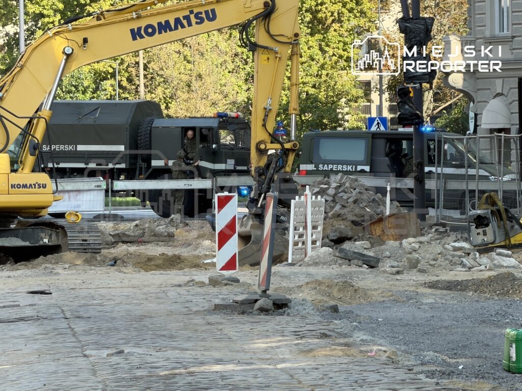 Koparka i wojskowe pojazdy saperskie przy ogrodzonym wykopie na rozkopanej ulicy, obok gruzu i znaków drogowych.