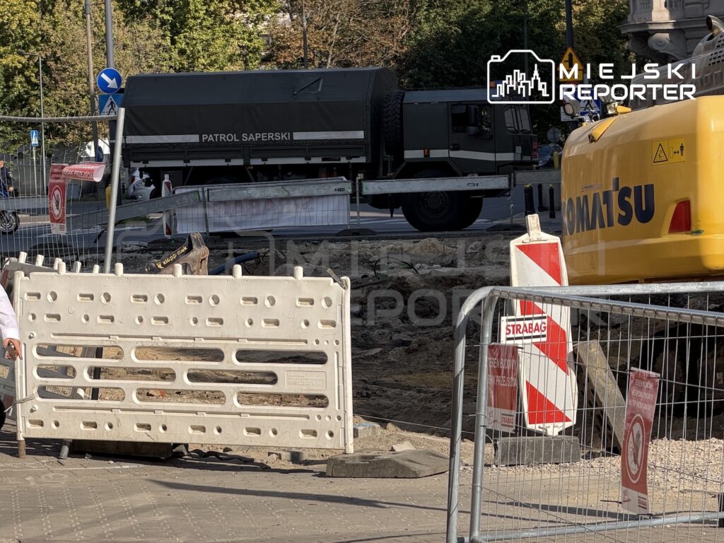 Samochód patrolu saperskiego za barierami i ogrodzeniem przy rozkopanej ulicy oraz żółtej koparce.