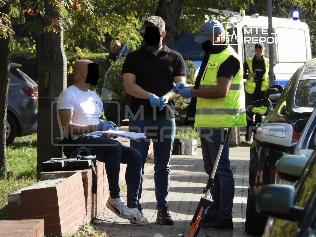 Ratownik w kamizelce odblaskowej udziela pomocy mężczyźnie w rękawiczkach, obok siedzi drugi mężczyzna na chodniku.