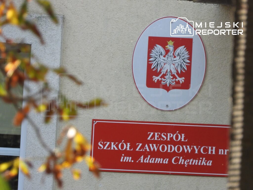 Tablica z godłem Polski i napisem „Zespół Szkół Zawodowych nr… im. Adama Chętnika” na elewacji budynku.
