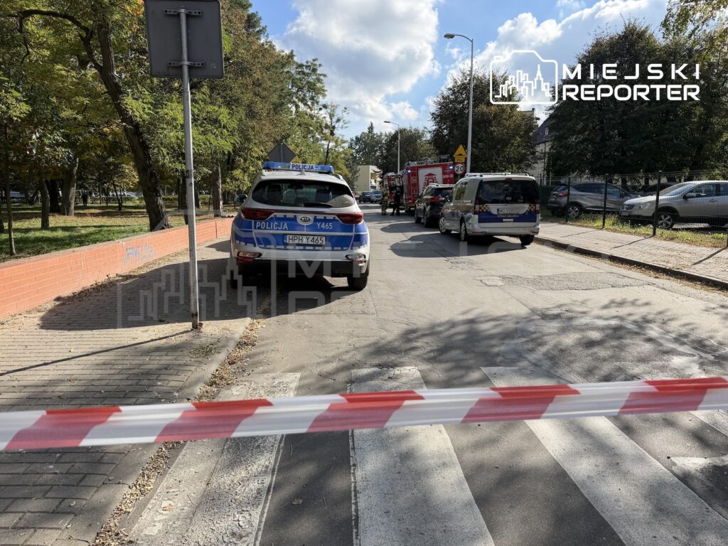 Radiowóz policyjny i pojazdy służb stoją na ulicy odgrodzonej biało-czerwoną taśmą.