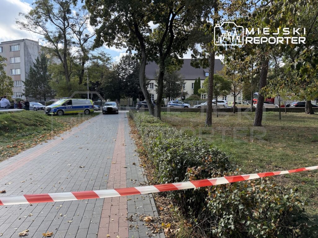 Taśma policyjna odgradza chodnik wśród drzew, a w tle stoi oznakowany samochód policji.