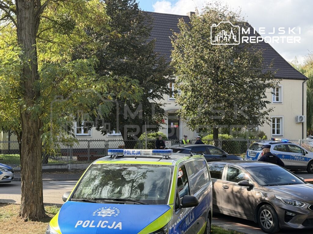Radiowozy policyjne zaparkowane przed budynkiem za ogrodzeniem, przy zadrzewionej ulicy.