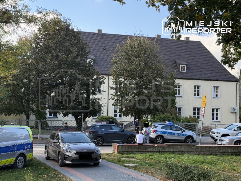 Radiowozy policyjne i samochody stoją przed jasnym, dwupiętrowym budynkiem otoczonym drzewami.