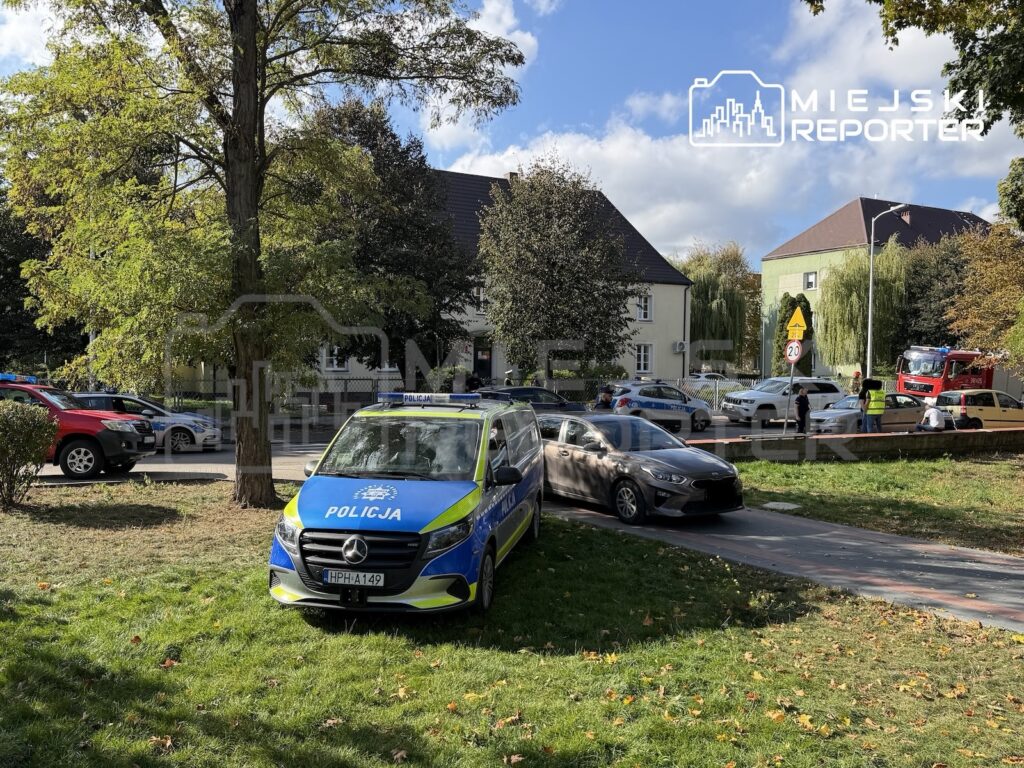 Radiowóz policji stoi przy ulicy przed budynkiem otoczonym taśmą, obok zaparkowanych samochodów i służb ratunkowych.