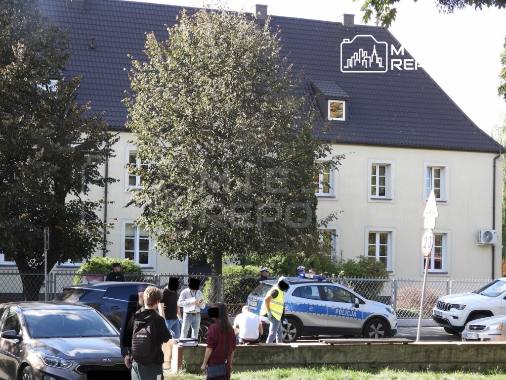 Policjanci i osoby postronne przed jasnym budynkiem; przy ogrodzeniu stoi niebiesko-biały radiowóz.