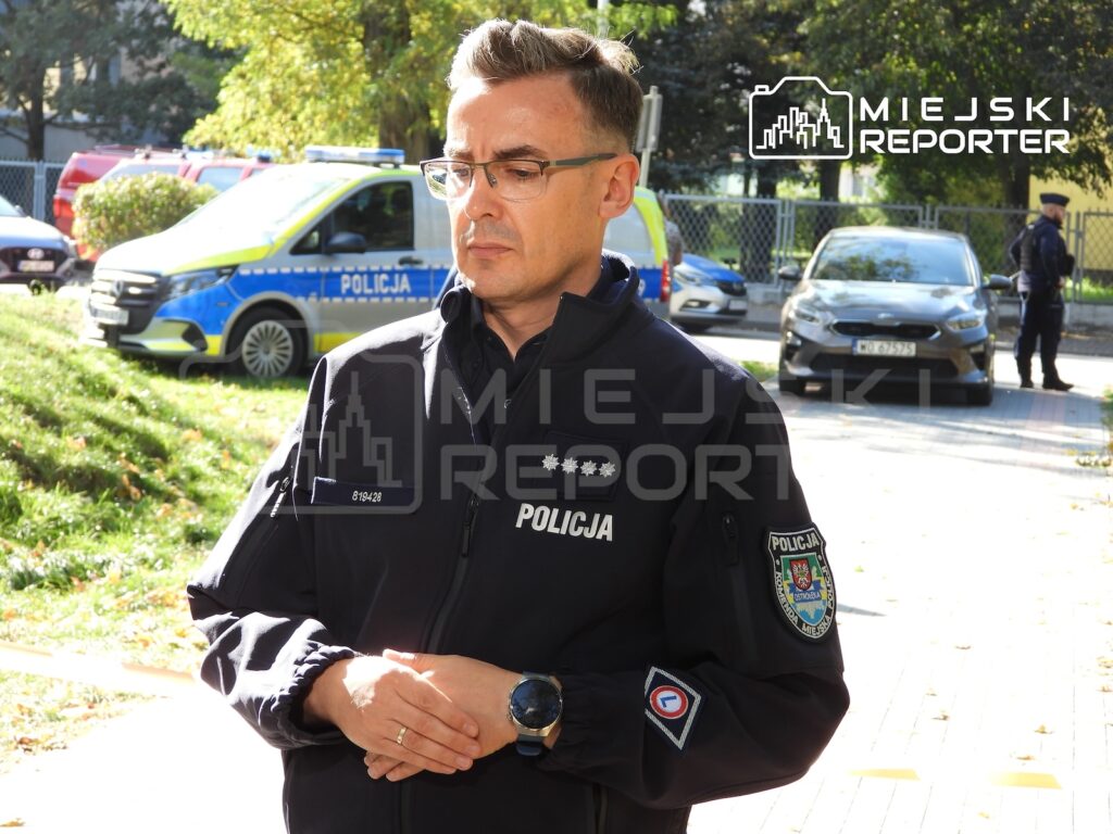 Policjant w mundurze stoi przed radiowozem i samochodem, obok ogrodzonego terenu z drzewami.