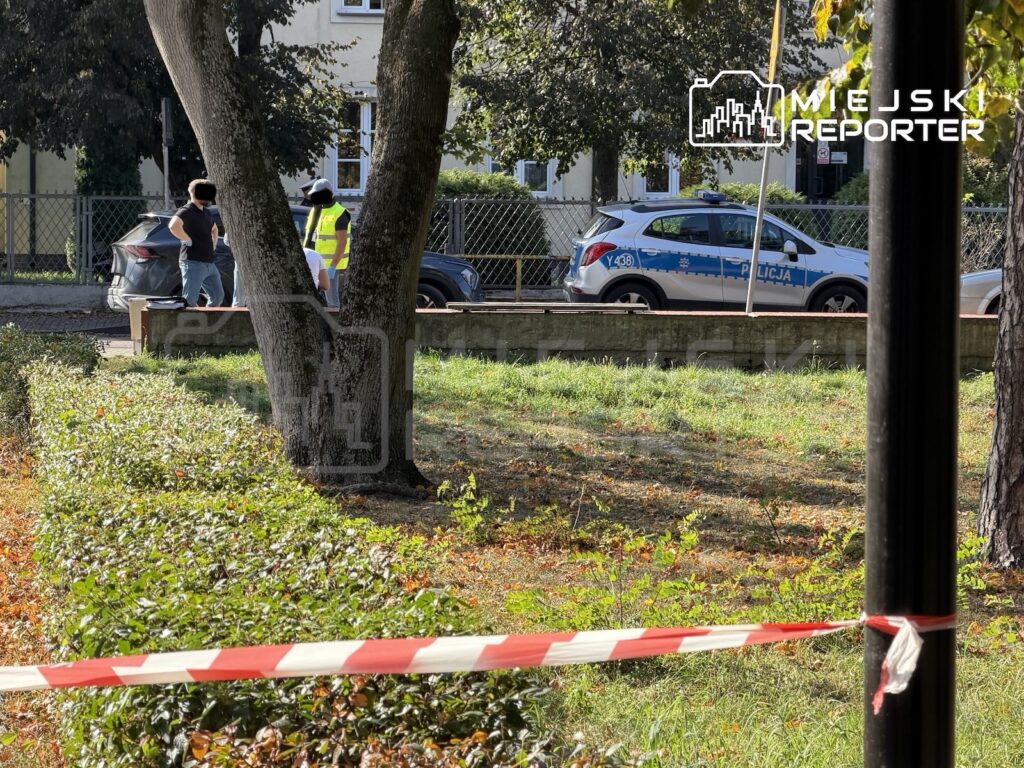 Radiowóz policyjny i funkcjonariusz za taśmą zabezpieczającą teren przy ogrodzeniu budynku.