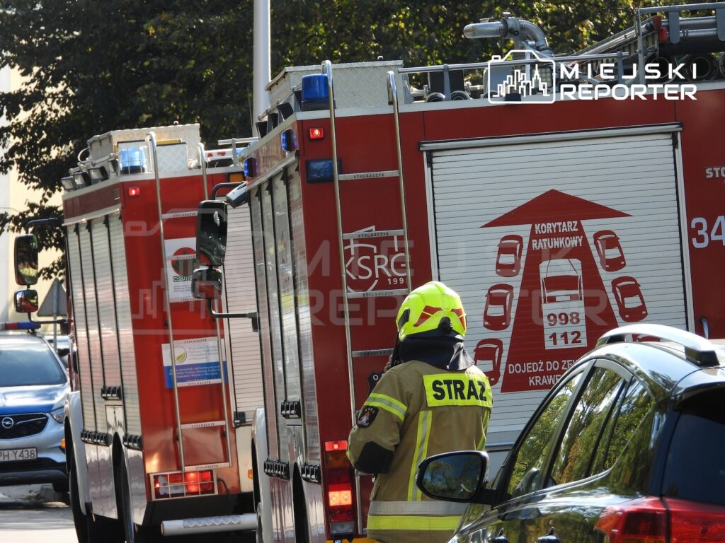 Strażak w hełmie stoi obok czerwonych wozów strażackich zaparkowanych przy ulicy między samochodami.