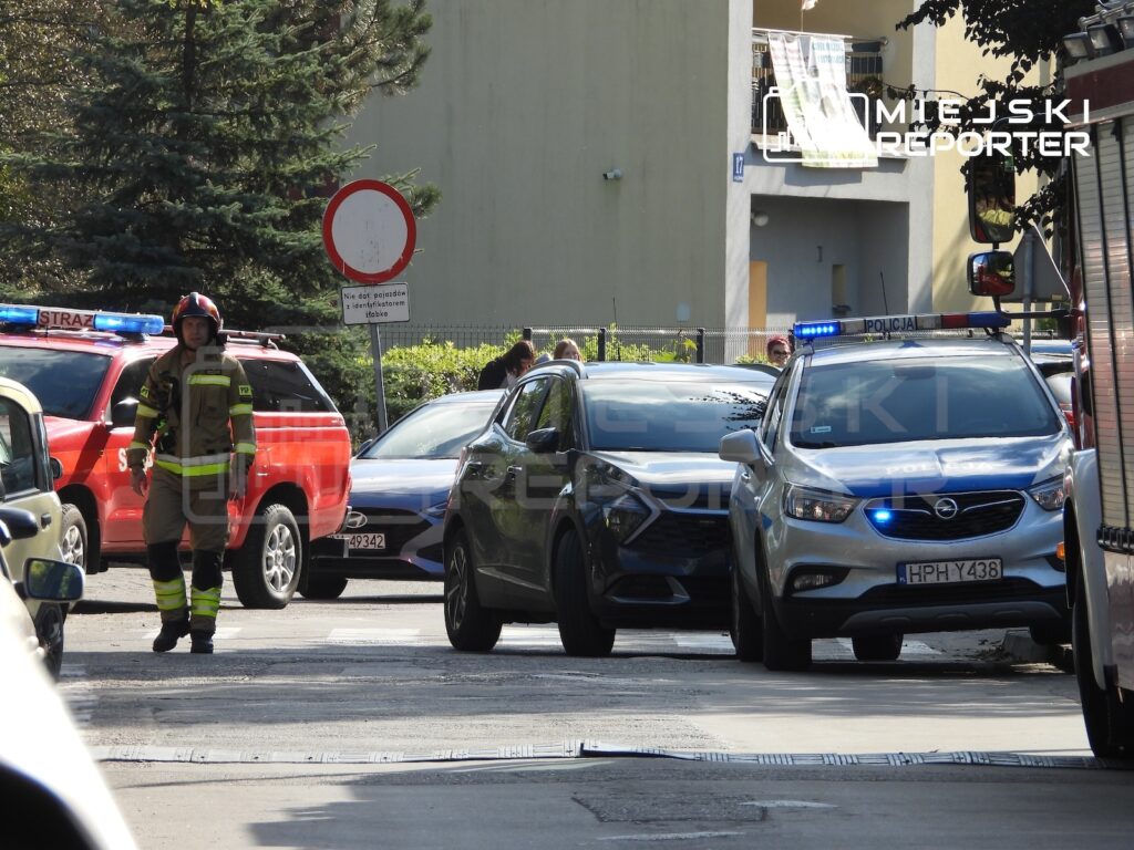 Strażak stoi obok wozu strażackiego, a radiowóz z włączonymi niebieskimi światłami stoi przy samochodzie osobowym przed