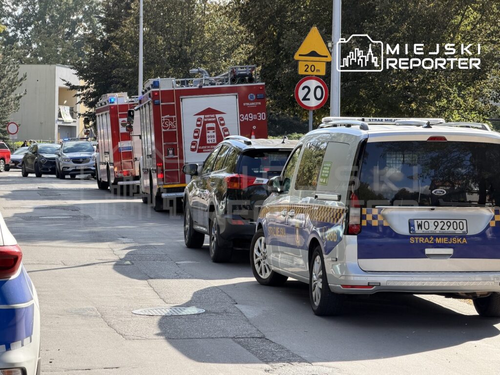Radiowóz straży miejskiej i wozy strażackie stoją na ulicy wśród zaparkowanych samochodów, przy znaku ograniczenia prędkości.