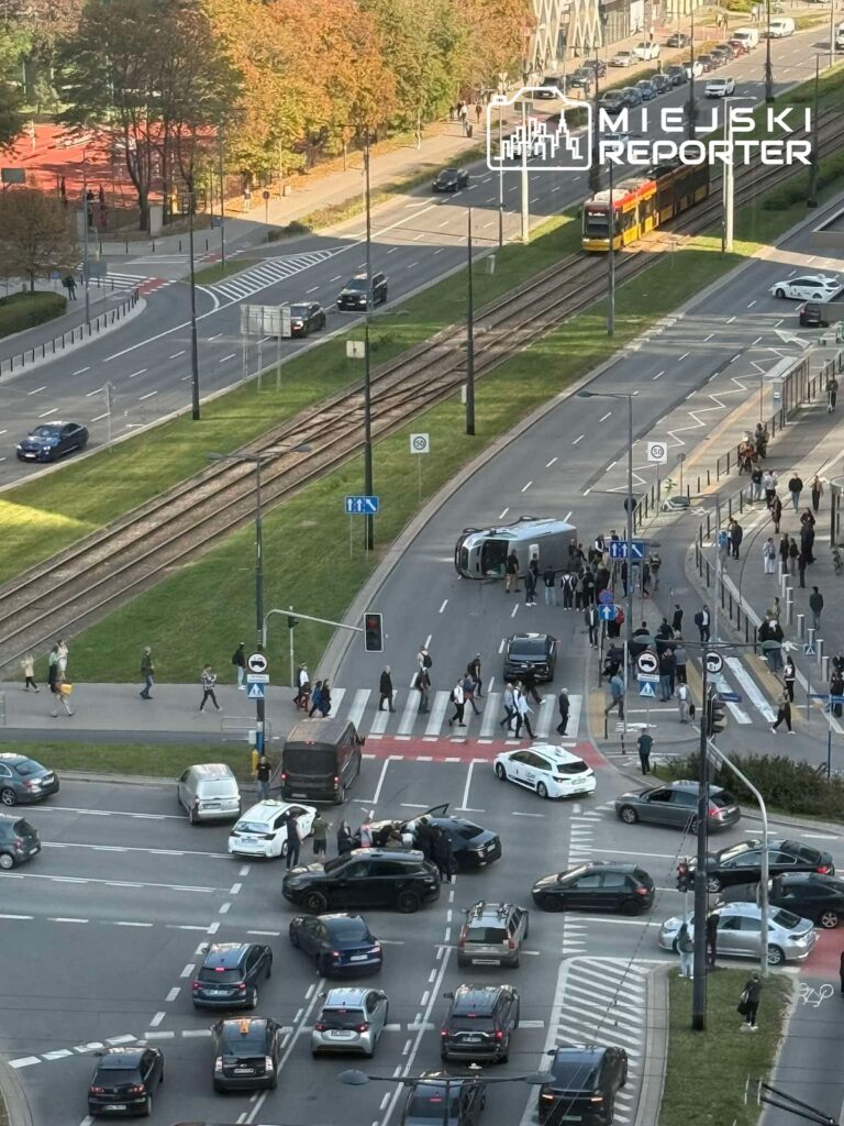 Przewrócony samochód dostawczy na skrzyżowaniu, wokół niego piesi i służby, a obok tramwaj na torach.