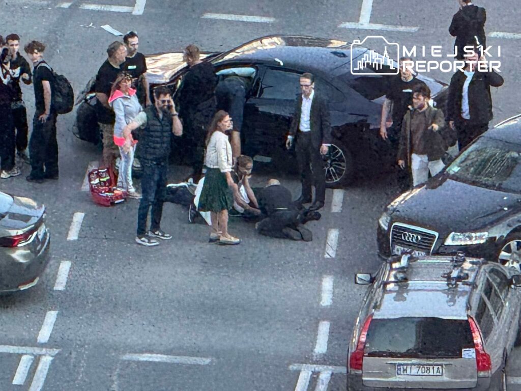 Grupa osób otacza leżącego człowieka między zaparkowanymi samochodami na parkingu.
