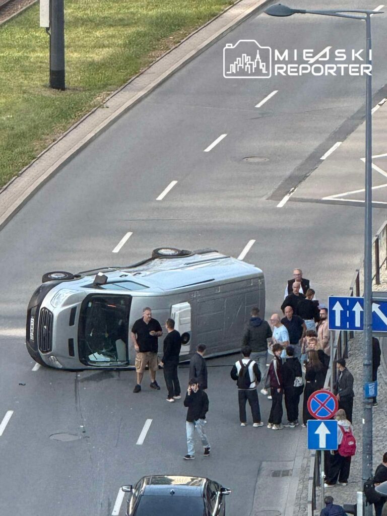 Przewrócony na bok bus blokuje jezdnię, a wokół pojazdu stoi grupa ludzi.