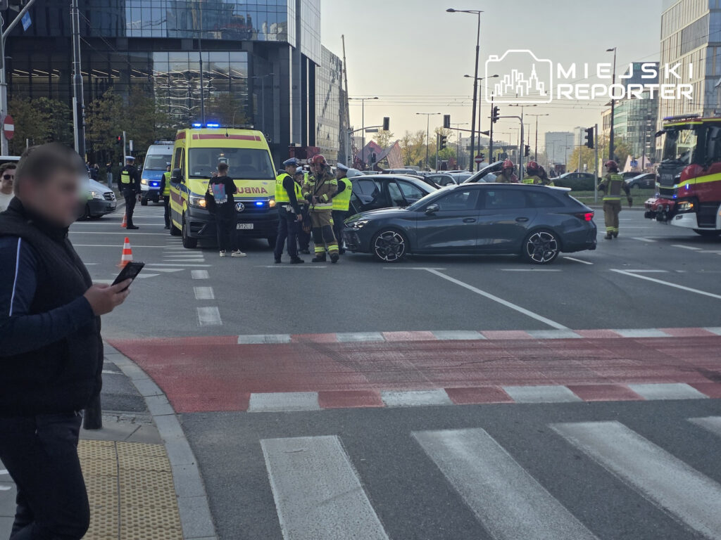 Ratownicy przy karetce udzielają pomocy przy zderzeniu dwóch samochodów na skrzyżowaniu; obok stoi wóz strażacki.