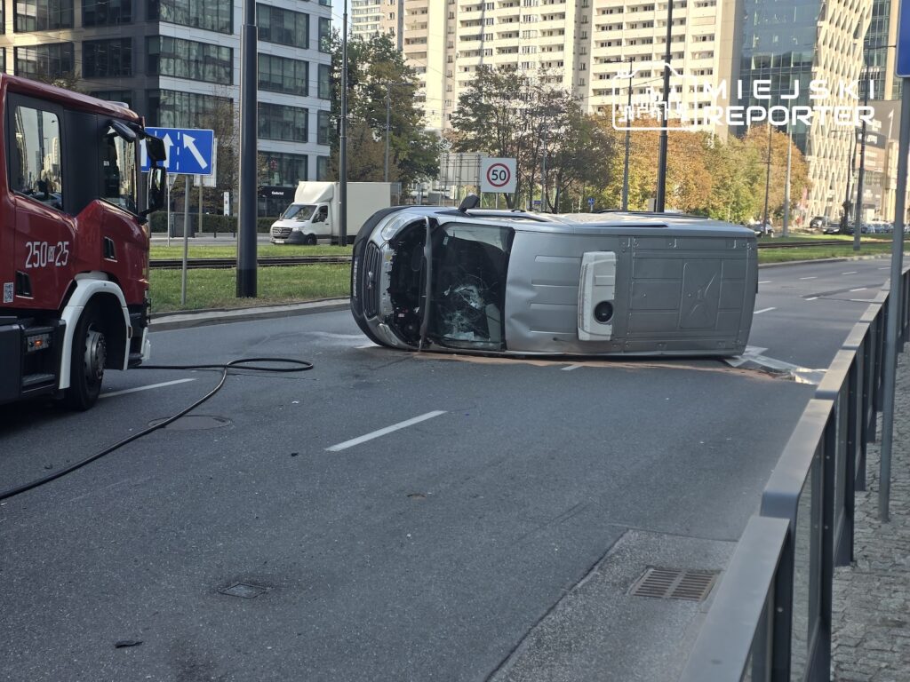 Przewrócony na bok szary samochód dostawczy blokuje pas ruchu; obok stoi czerwony wóz strażacki.