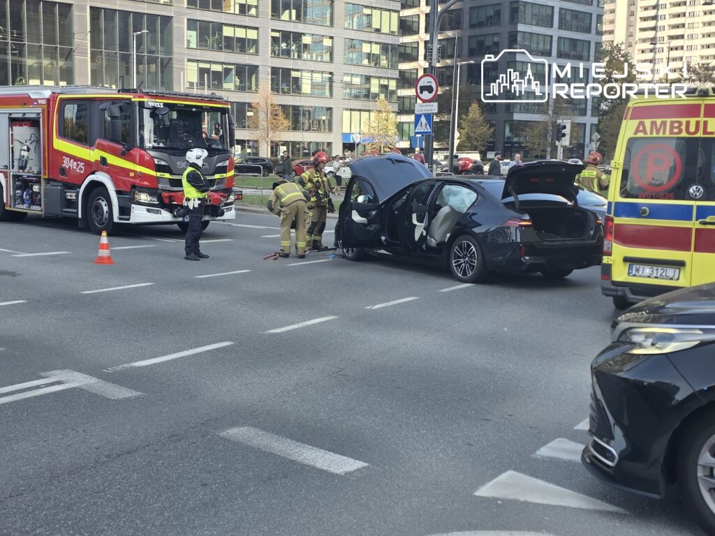 Strażacy i ratownicy przy rozbitym samochodzie osobowym między wozem strażackim a karetką na miejskiej ulicy.