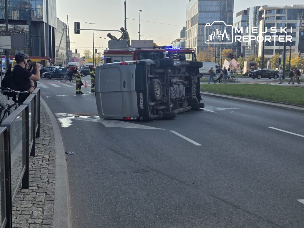 Przewrócony na bok bus blokuje jezdnię, a obok pracują strażacy i stoi wóz strażacki.