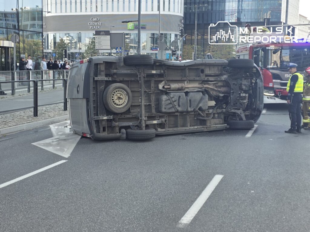 Przewrócony na bok samochód dostawczy blokuje pas jezdni przed przeszklonym budynkiem; obok stoi funkcjonariusz w