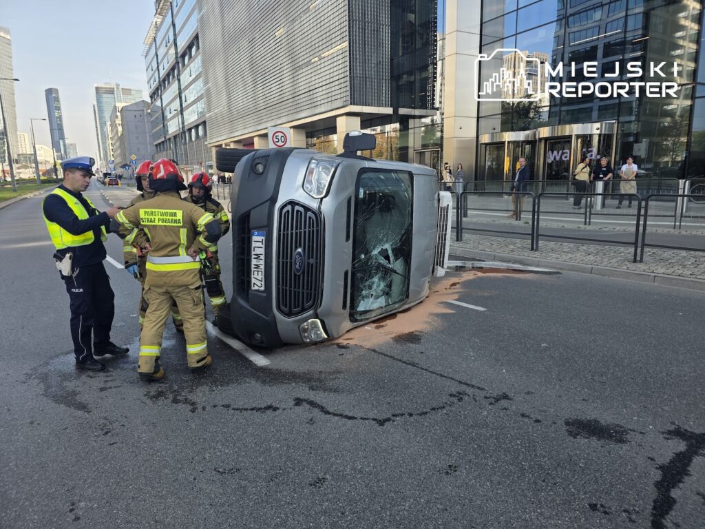 Przewrócony na bok samochód dostawczy na ulicy, obok policjant i strażacy w odblaskowych uniformach.