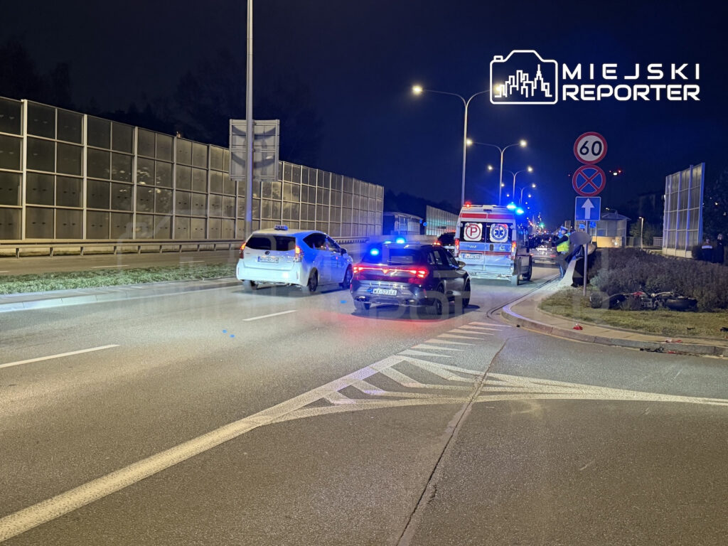 Karetka i samochody stoją na nocnej drodze przy barierach dźwiękochłonnych, a niebieskie światła pojazdu służb migają.