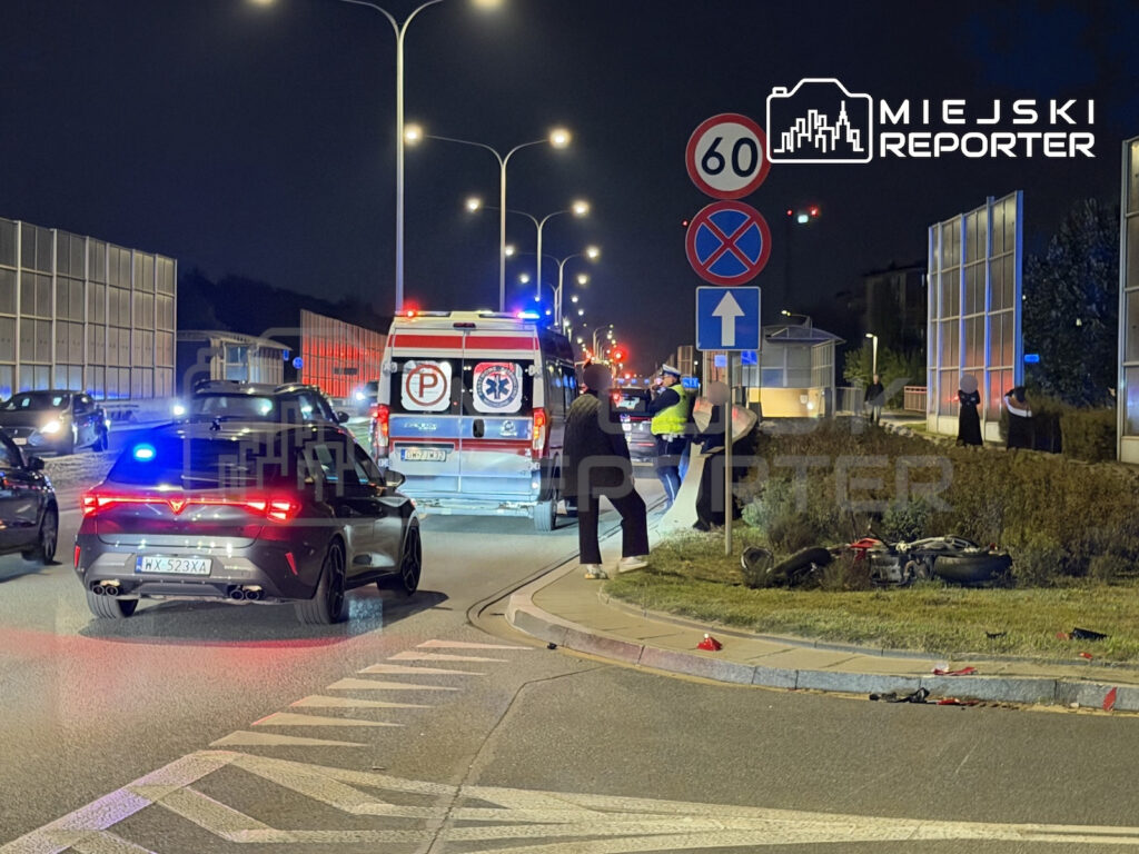 Karetka z włączonymi światłami stoi na nocnej drodze, a służby zabezpieczają miejsce zdarzenia przy skrzyżowaniu.