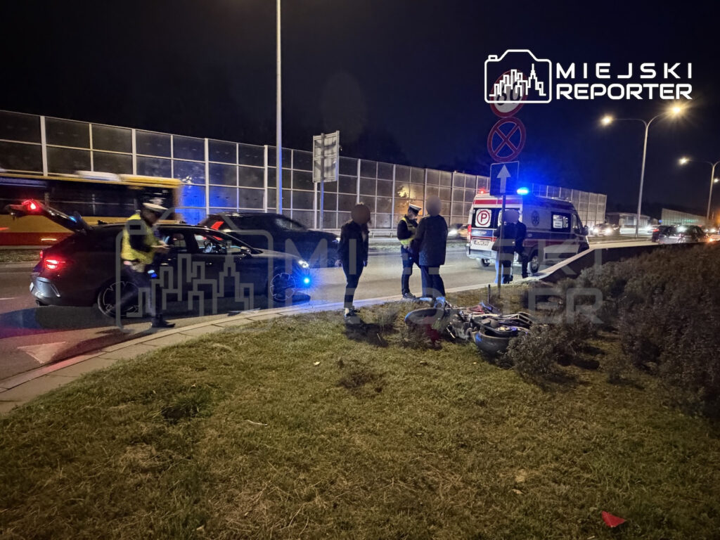 Służby i świadkowie przy rozbitym rowerze obok karetki i samochodów na nocnej ulicy.