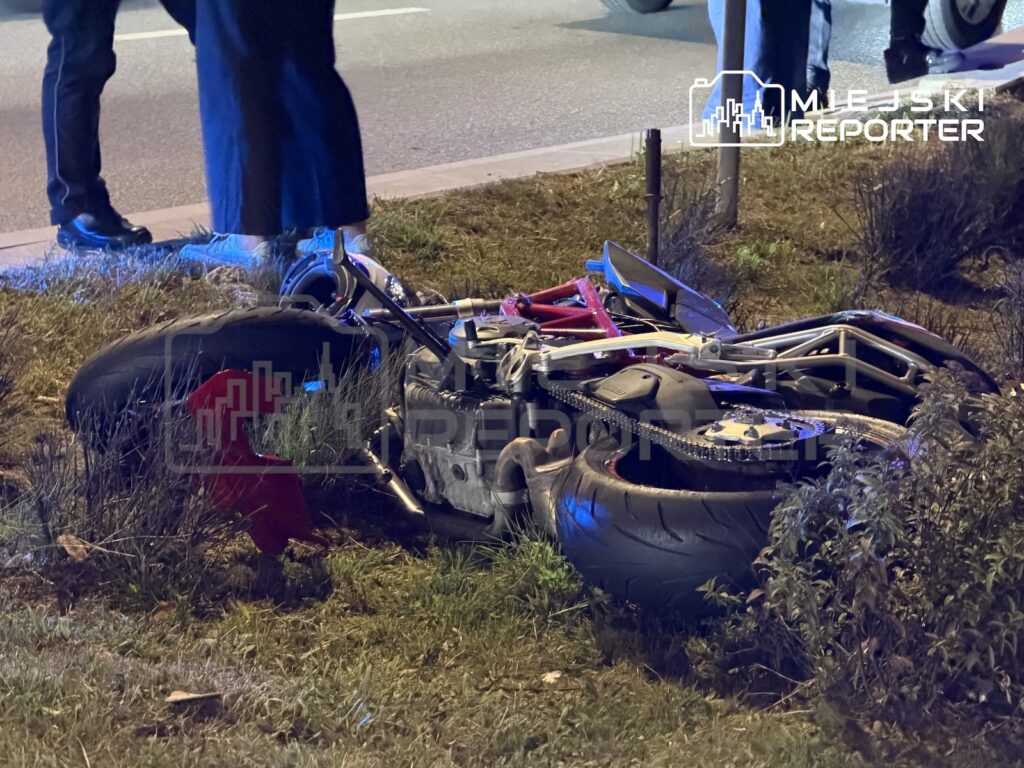Przewrócony motocykl leży na trawniku przy drodze, obok stoją osoby w mundurach.