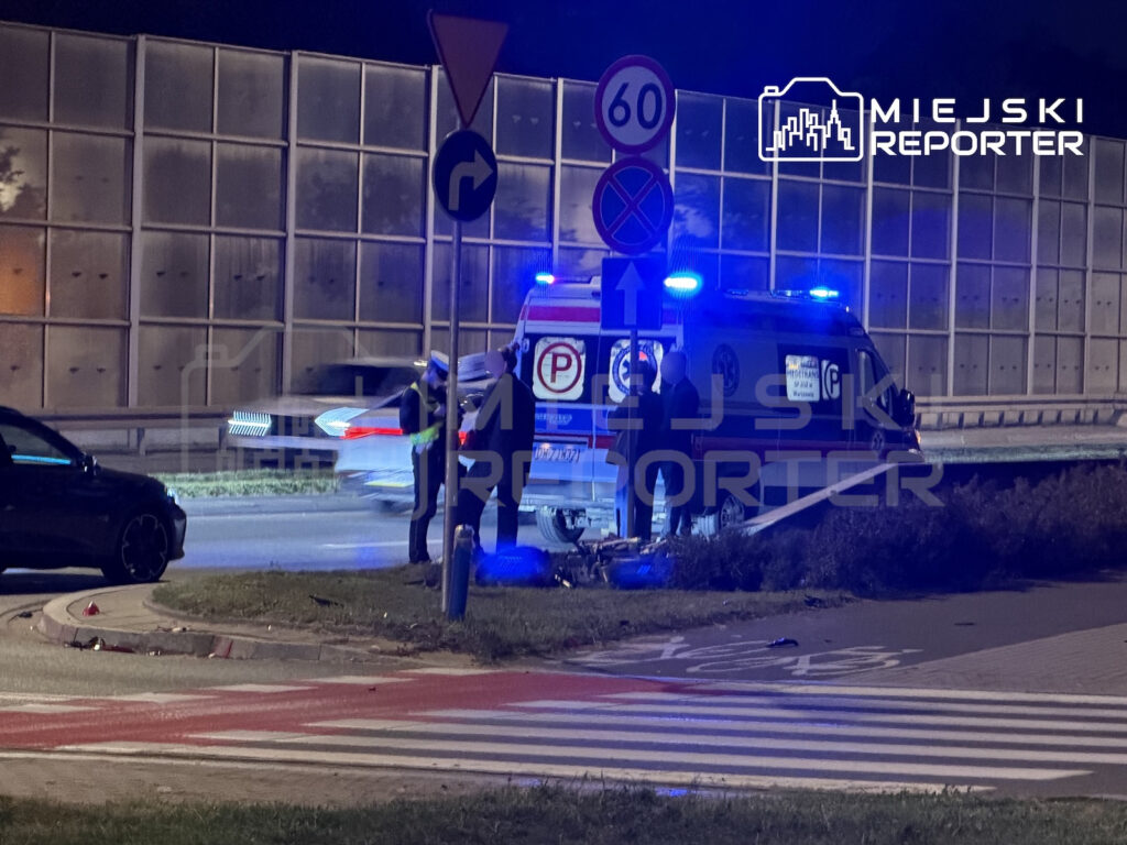 Karetka z włączonymi niebieskimi sygnałami stoi nocą przy przejściu dla pieszych, obok pracują służby ratunkowe.