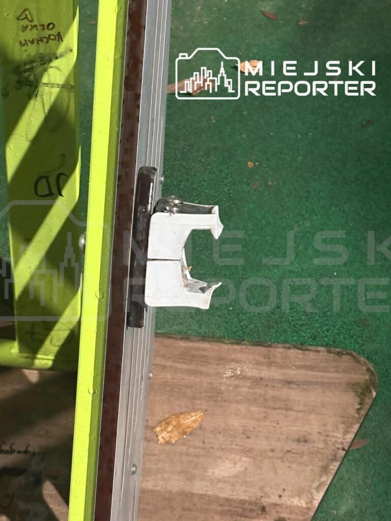 Zbliżenie na metalowy uchwyt przy zielonej konstrukcji placu zabaw i zużytą drewnianą platformę.