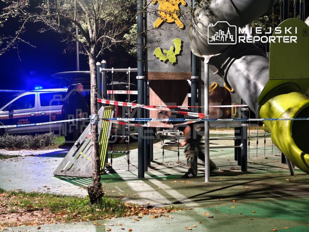 Taśma policyjna odgradza konstrukcję placu zabaw, obok stoi oznakowany samochód straży miejskiej z włączonymi światłami.