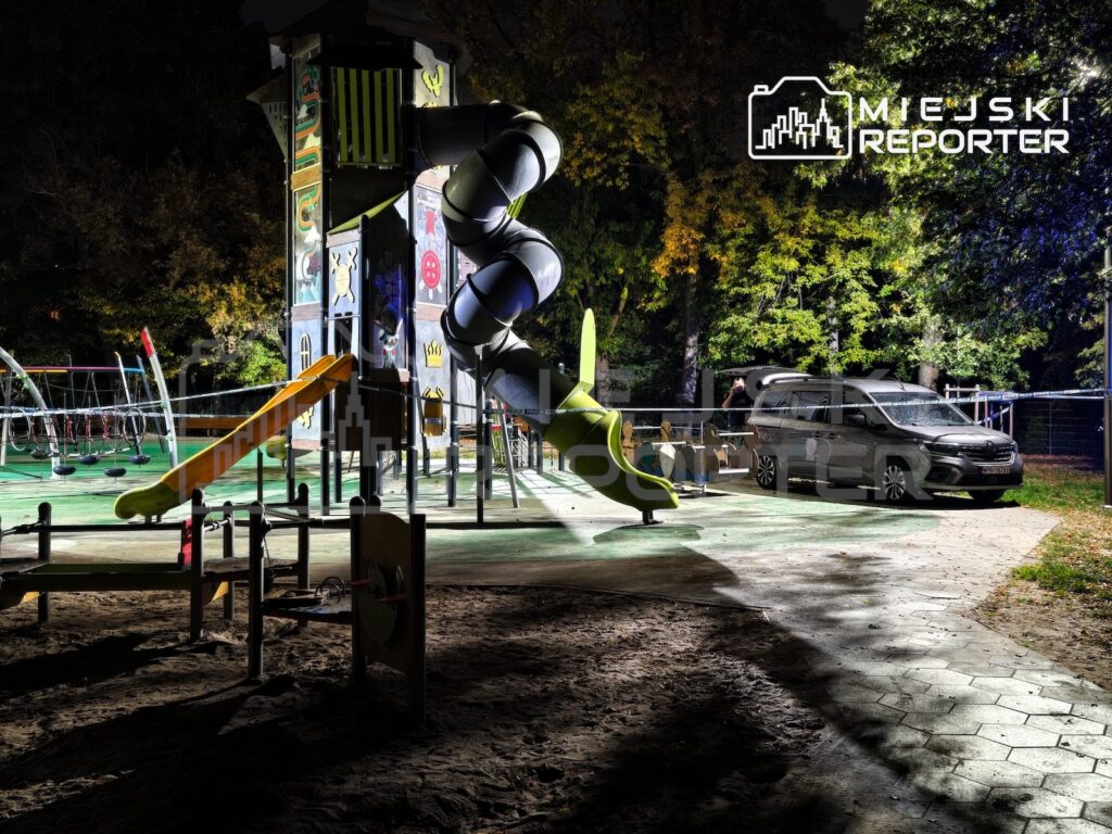 Oświetlony plac zabaw z wysoką konstrukcją, spiralną zjeżdżalnią i huśtawkami, nocą wśród drzew.