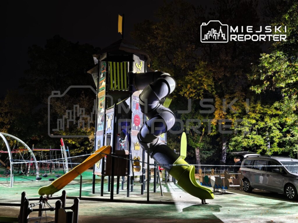 Oświetlony plac zabaw nocą z wysoką konstrukcją, krętą zjeżdżalnią i zaparkowanym obok samochodem