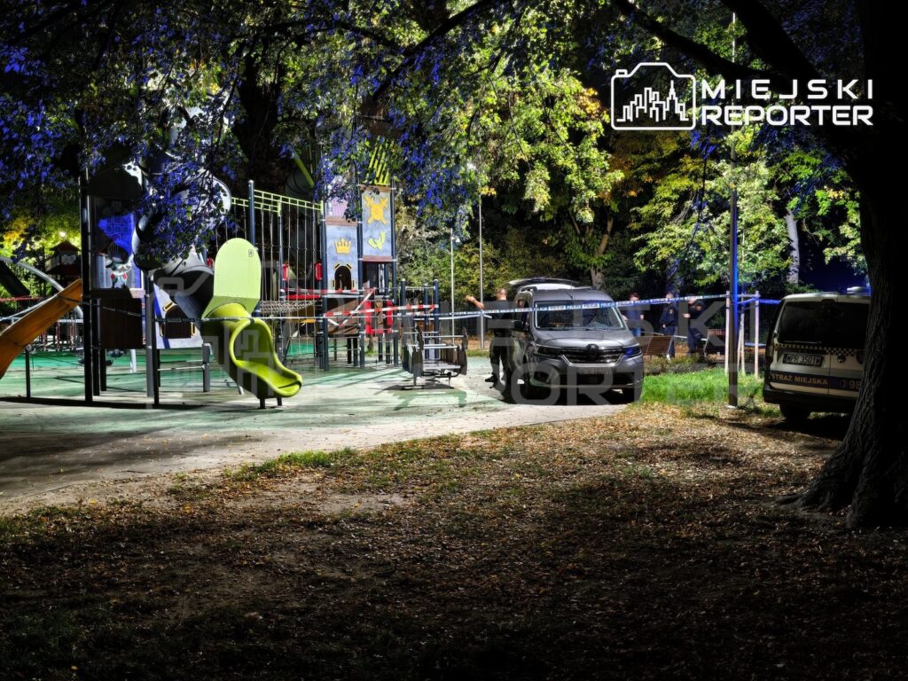 Oświetlony plac zabaw z zieloną zjeżdżalnią, odgrodzony taśmą, obok stoją samochody służb ratunkowych.