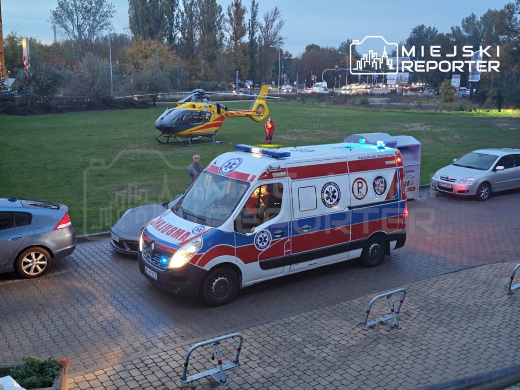 Karetka pogotowia stoi na parkingu, a żółty śmigłowiec ratunkowy ląduje na pobliskim trawniku.