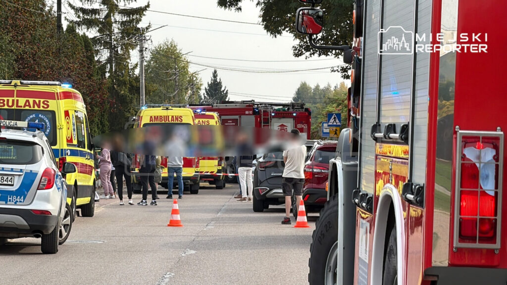 Ambulanse, radiowozy i wóz strażacki stoją na ulicy, gdzie służby zabezpieczają miejsce zdarzenia pachołkami.