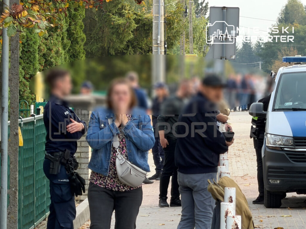 Zamaskowani ludzie stoją na chodniku przy ogrodzeniu, obok funkcjonariuszy i białego busa policyjnego.