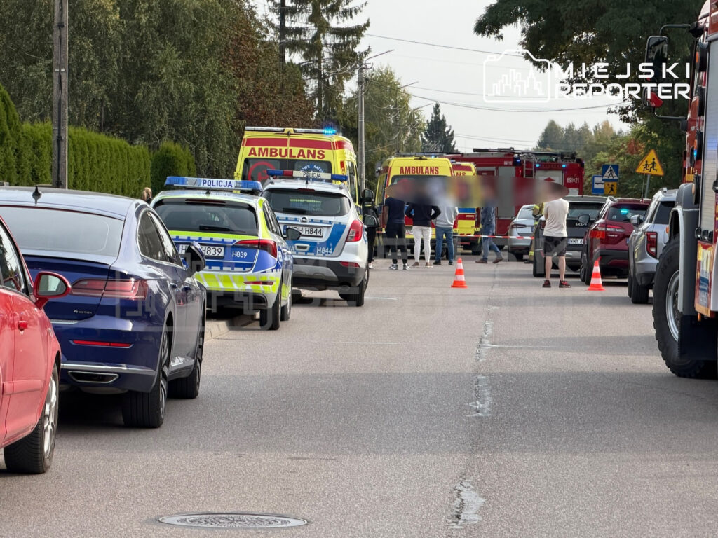 Karetka pogotowia, radiowozy i wozy strażackie stoją na ulicy, gdzie służby zabezpieczają miejsce zdarzenia.