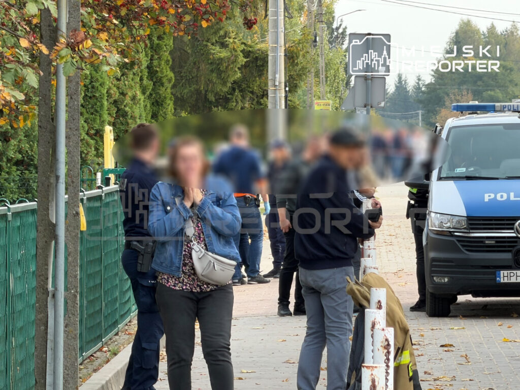 Policjanci i cywile stoją na chodniku obok policyjnego radiowozu przy ogrodzeniu szkoły.