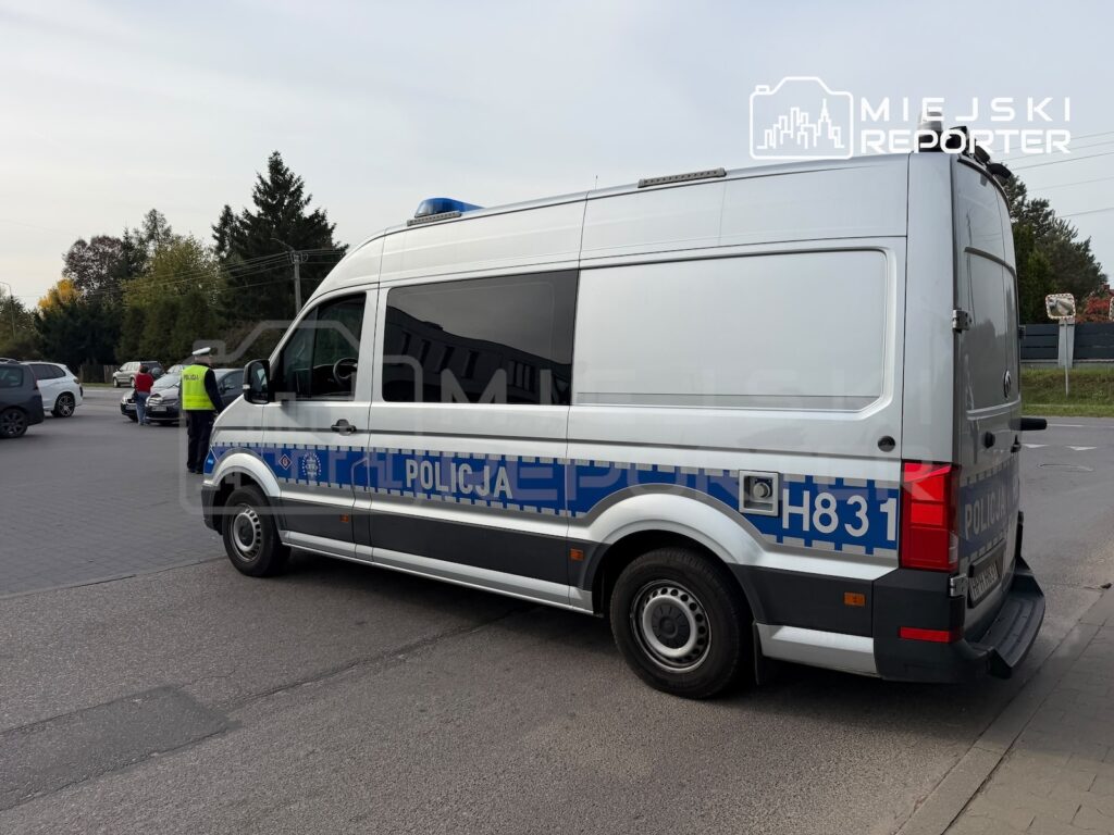 Policyjny bus z napisem „POLICJA” stoi na parkingu, obok funkcjonariusz w kamizelce odblaskowej.