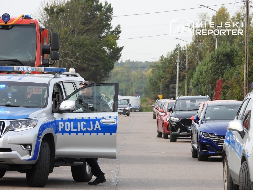 Radiowóz z napisem „POLICJA” z otwartymi drzwiami i włączonymi sygnałami świetlnymi na ulicy, obok wozu strażackiego i