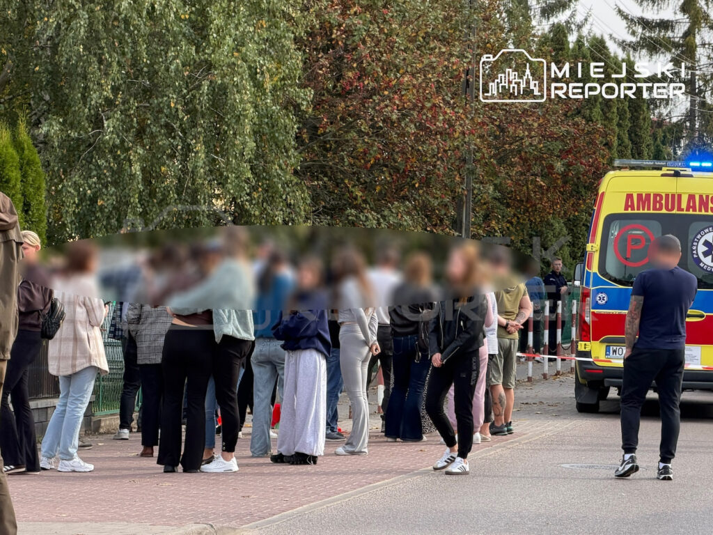 Grupa osób stoi przy zaparkowanej karetce pogotowia na zadrzewionym chodniku; nad pojazdem błyska niebieski sygnał świetlny.