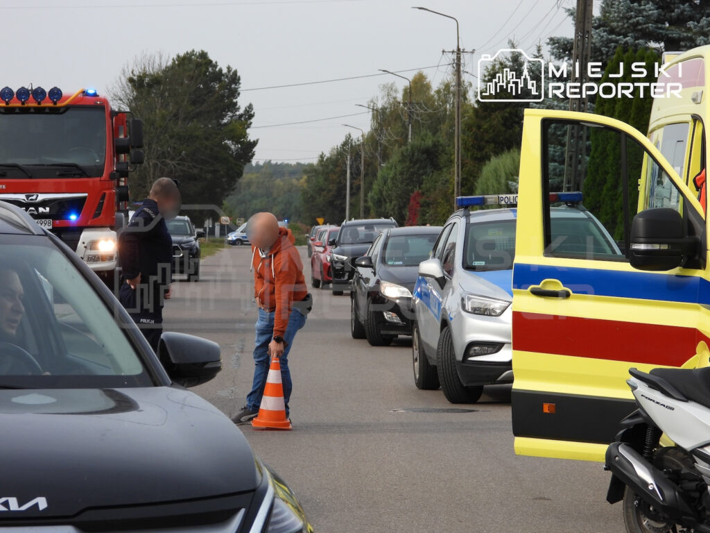 Policjant i ratownik zabezpieczają miejsce zdarzenia na drodze, obok radiowozu, karetki i wozu strażackiego.